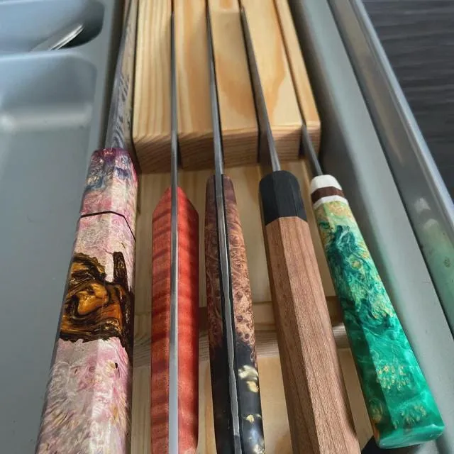 Tiroir ouvert : cinq couteaux aux manches colorés, rangés à plat dans un bloc de bois à fentes.